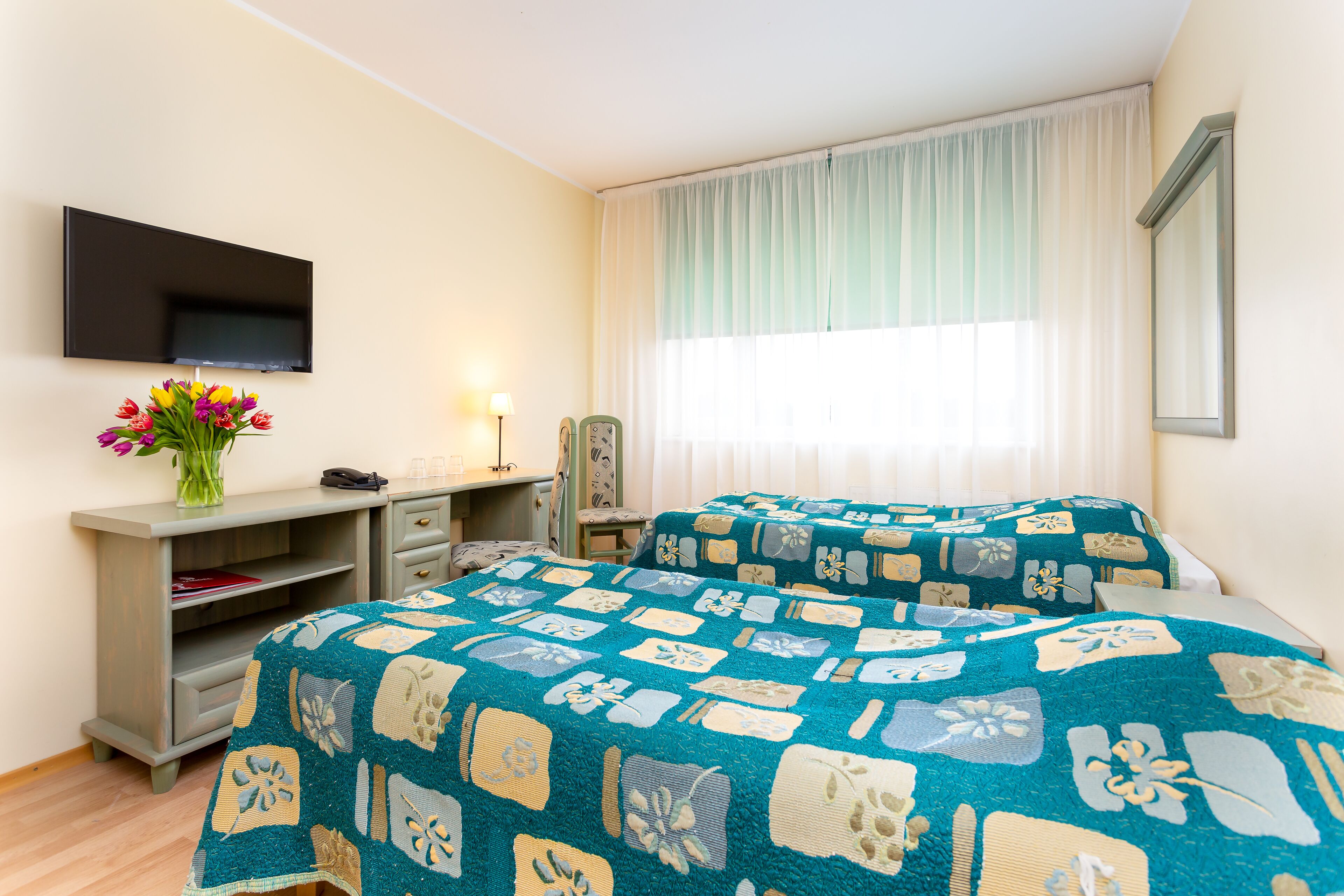 A hotel room with two beds, a TV, a desk with a lamp, and a window with curtains.