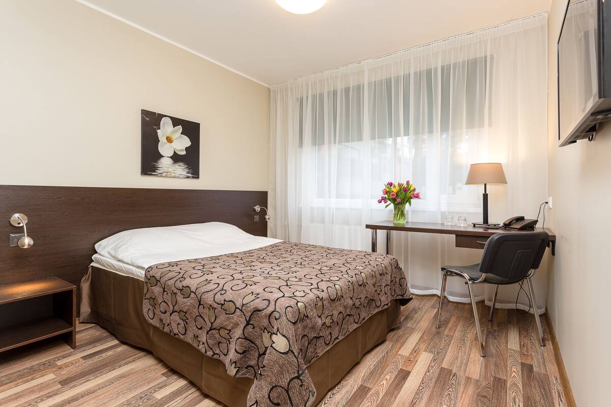 A hotel room with a bed, a desk with a chair, a lamp, a flower vase, and a picture on the wall.