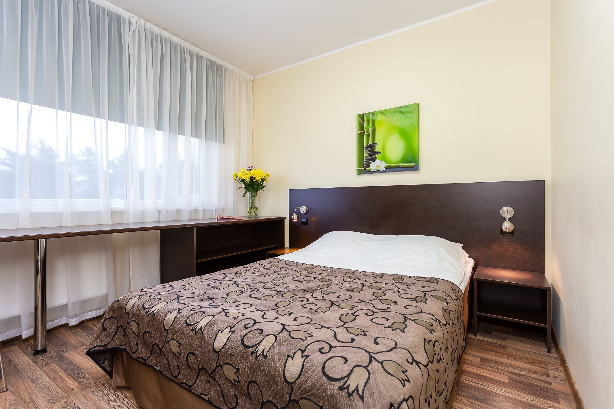 A hotel room with a bed, a desk, a chair, a nightstand, a lamp, and a painting on the wall.