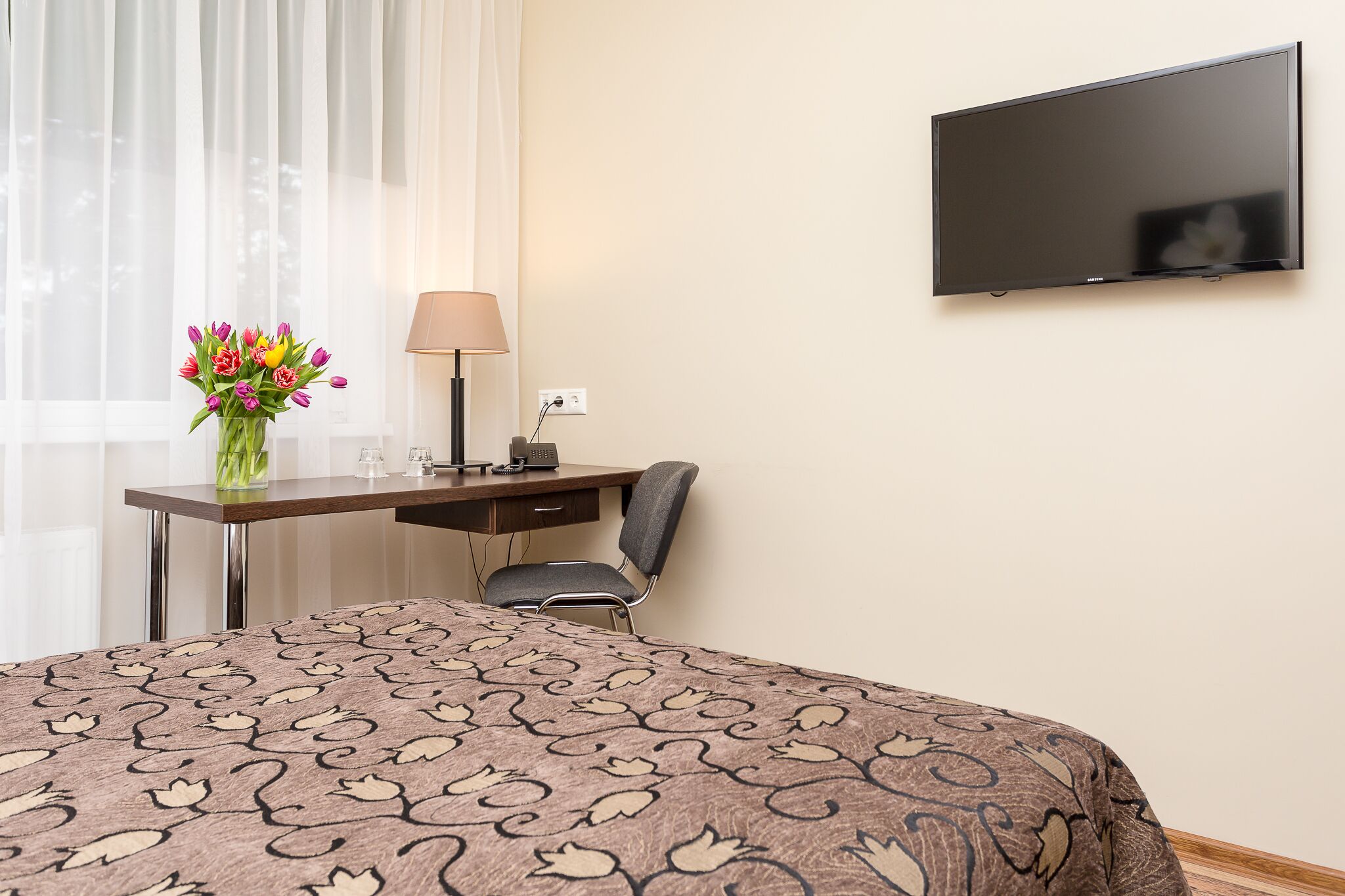 Habitación de hotel con cama, escritorio con lámpara, silla, televisor montado en la pared y ventana con cortinas traslúcidas.