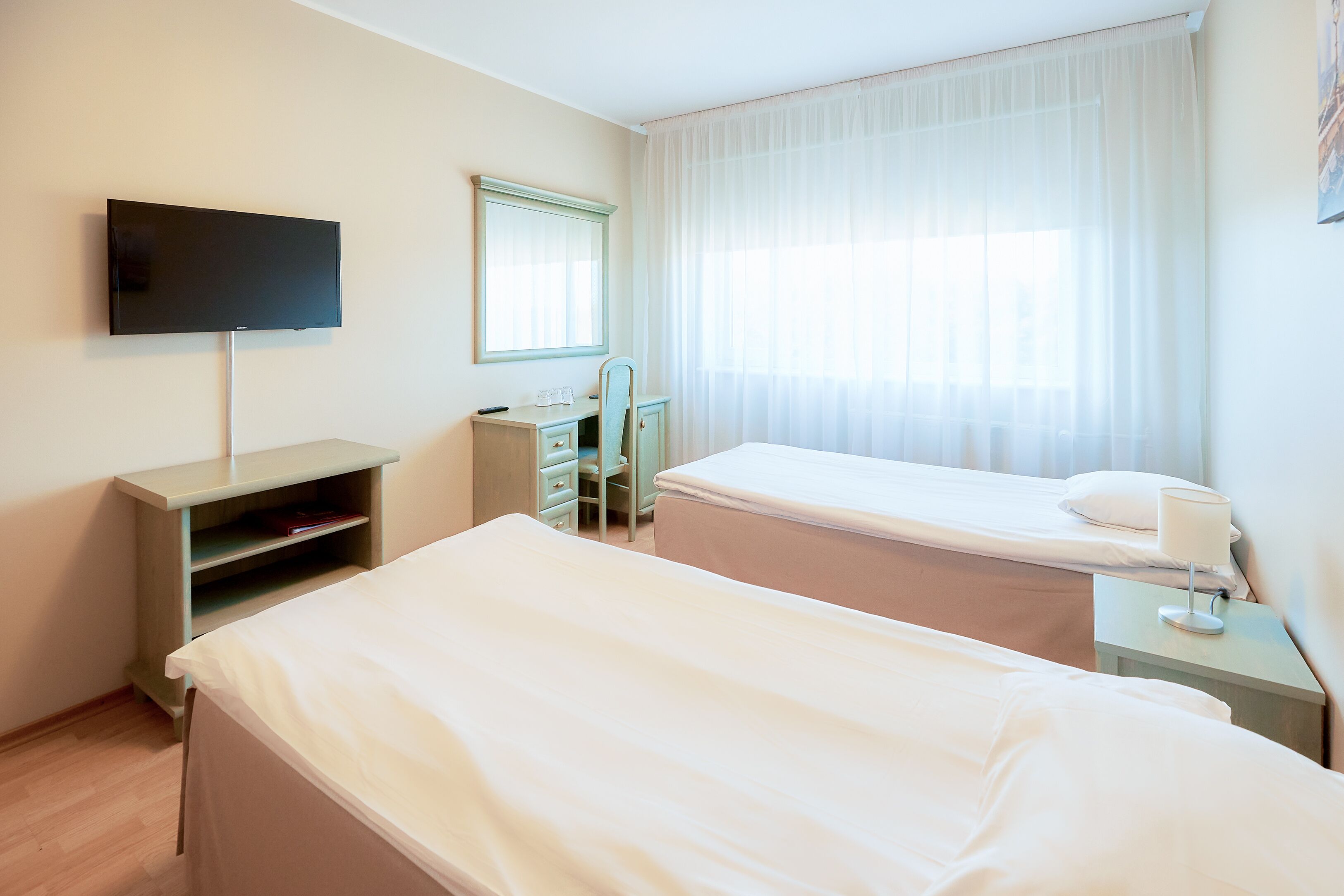 A hotel room with two beds, a TV, a desk, and a window with sheer curtains.