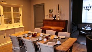 A dining area with a wooden table set for six, surrounded by wicker chairs. A piano is positioned against the wall, and a cabinet with glass doors is visible to the left.