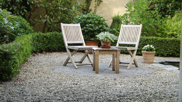 A garden patio with two wooden chairs and a table, surrounded by gravel and potted plants.