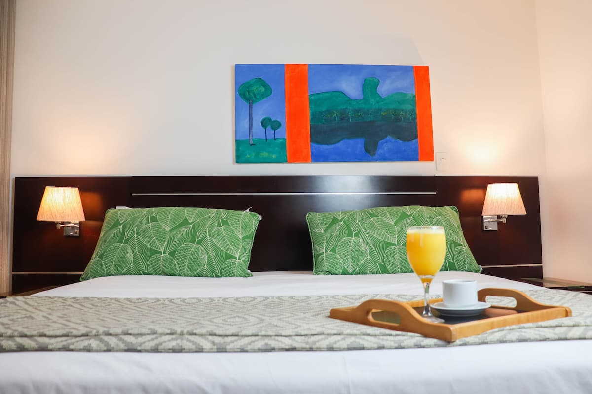 A neatly made bed with green patterned pillows, a wooden tray with a glass of orange juice, and a painting above the bed.