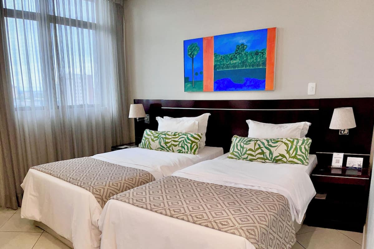A hotel room with two beds, a painting on the wall, and a window with curtains.