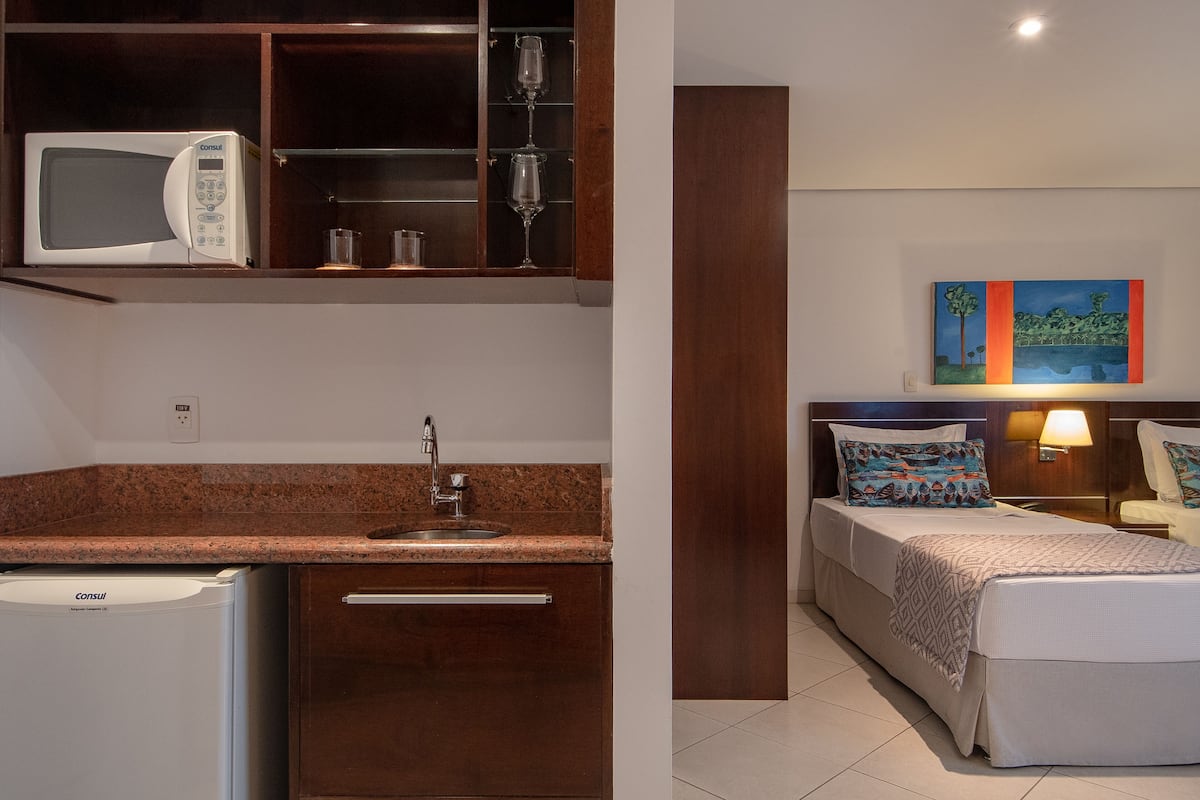 A compact hotel room with a kitchenette, a bed with patterned bedding, a bedside table with a lamp, and a painting on the wall.