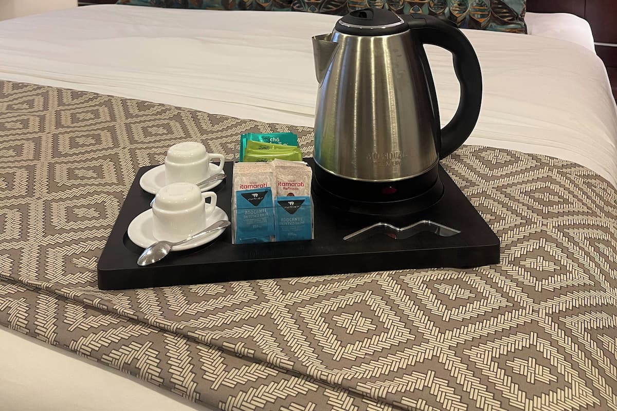 A neatly made bed with a patterned bedspread, a tray with tea bags, a teapot, and a cup and saucer on a black tray.