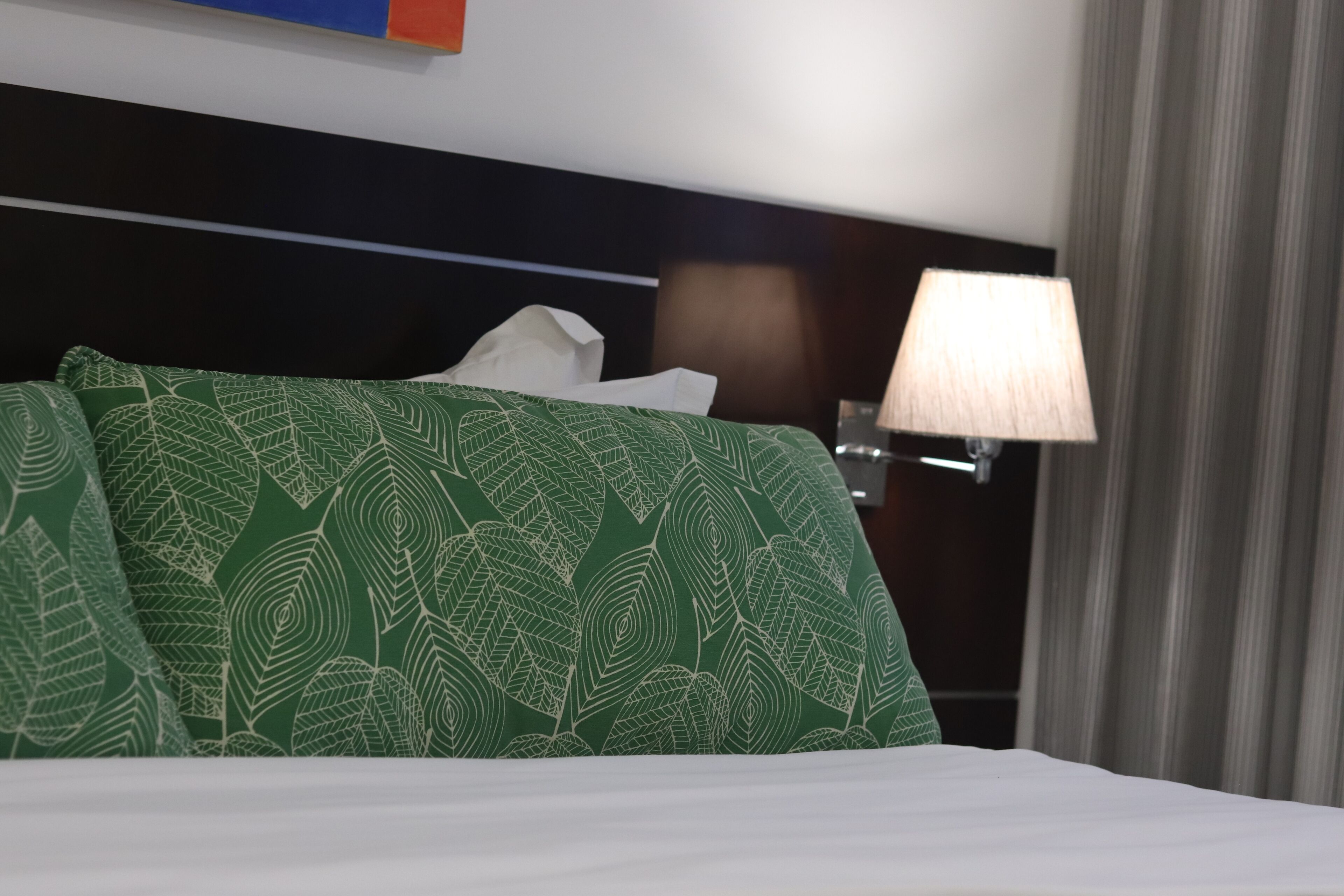 Ein Hotelzimmer mit einem ordentlich bezogenen Bett, einer grün gemusterten Tagesdecke, einem weißen Kissen, einer Nachttischlampe und einem Fenster mit Vorhängen.