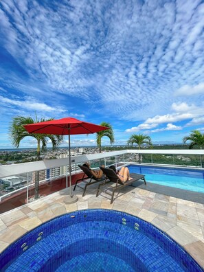 Zona de piscina en la azotea con sillones de descanso y un paraguas rojo, con vistas a un paisaje urbano.