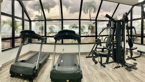 A gym with treadmills, weight machines, and a large window offering a view of palm trees and a cloudy sky. - Manaus Hotéis - Millennium (Manaus)