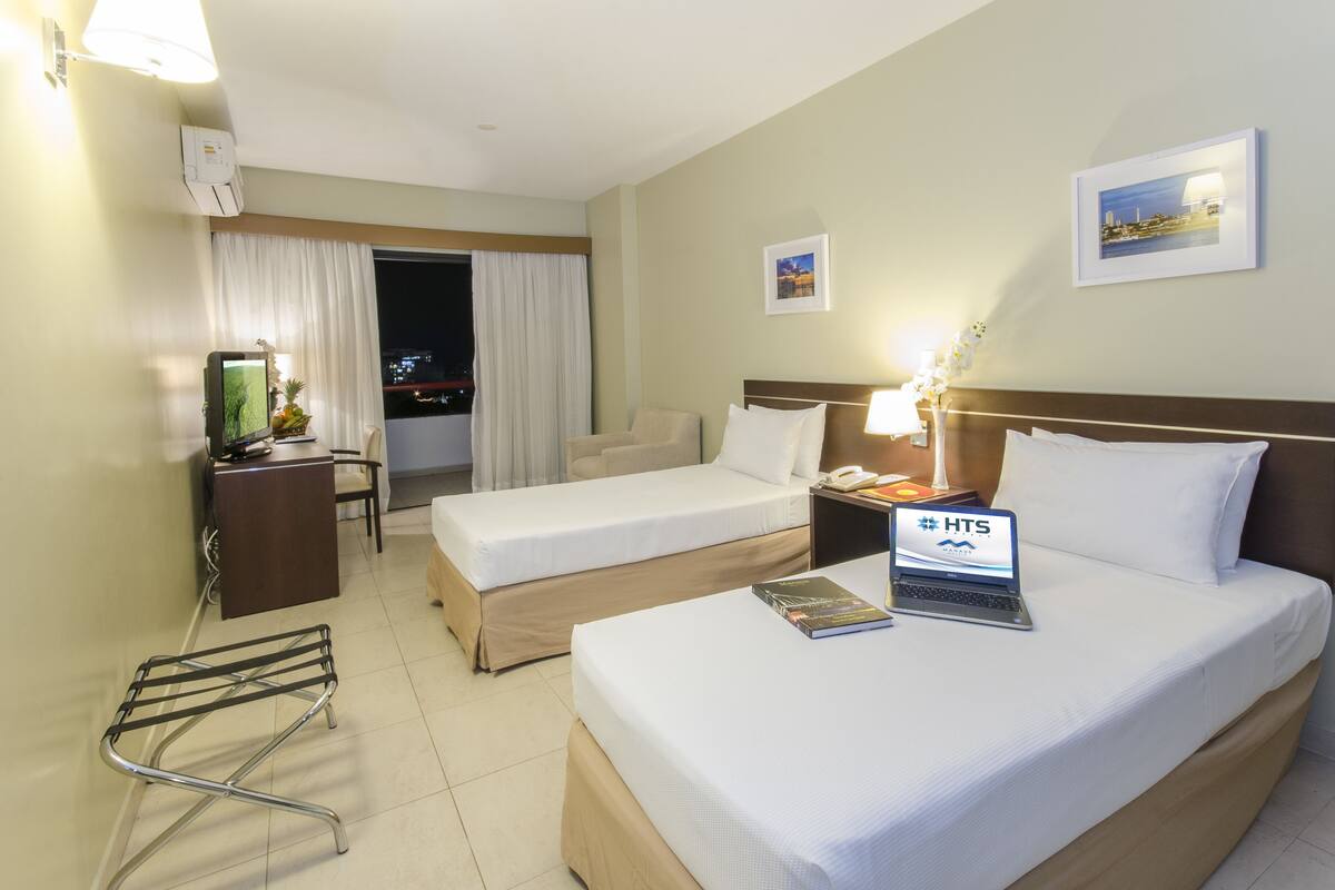 A hotel room with two beds, a desk with a computer, a chair, a small table with a lamp, and a window with curtains.