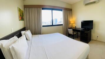 Una habitación de hotel con una cama grande, un escritorio con televisor, aire acondicionado y una ventana con cortinas.