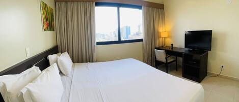 A hotel room with a large bed, a desk with a TV, an air conditioner, and a window with curtains.