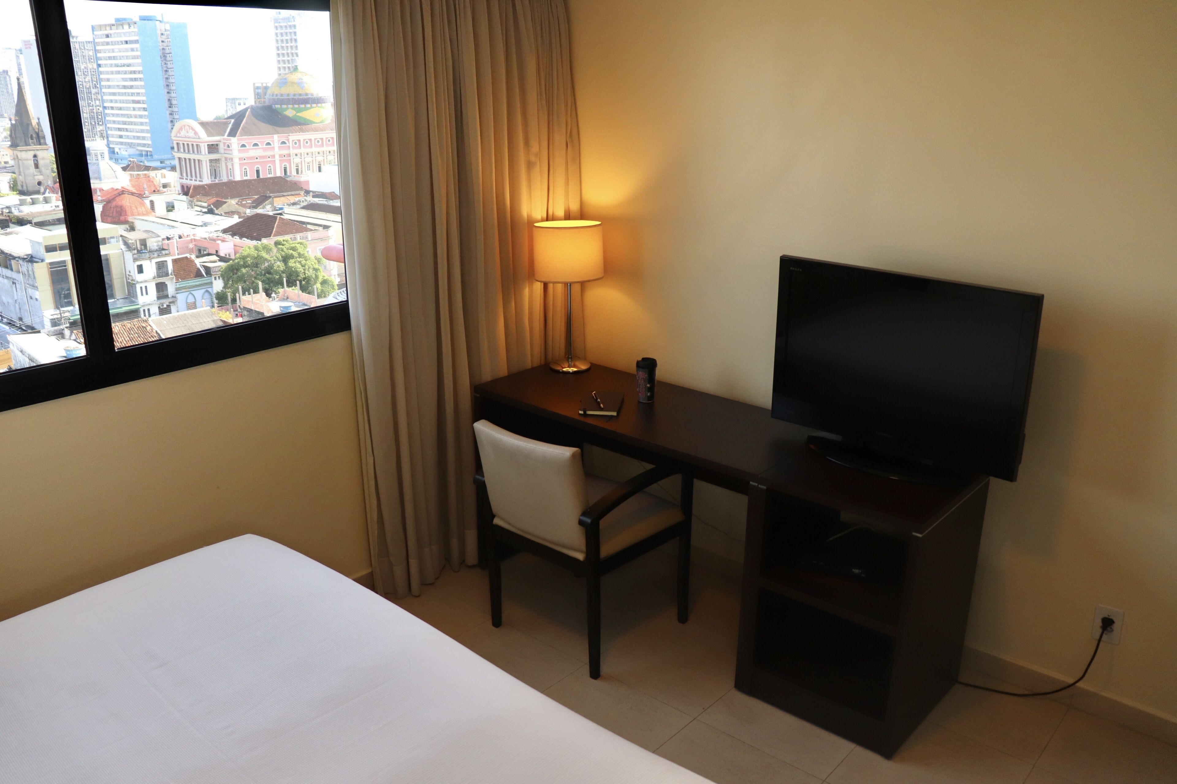 Quarto de hotel com cama, escrivaninha com televisão, cadeira, luminária e vista para a cidade através de uma janela.