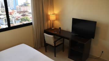 A hotel room with a bed, a desk with a TV, a chair, a lamp, and a view of a cityscape through a window.