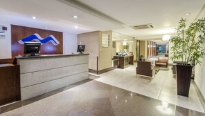 A hotel reception area with a reception desk, a digital screen, a reception counter, a seating area with chairs, and a large potted plant. - Hotel Saint Paul (Manaus)