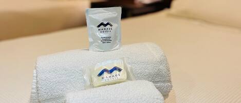 A neatly made bed with white towels and a small sachet of hand soap labeled "MANAUS HOTELS".