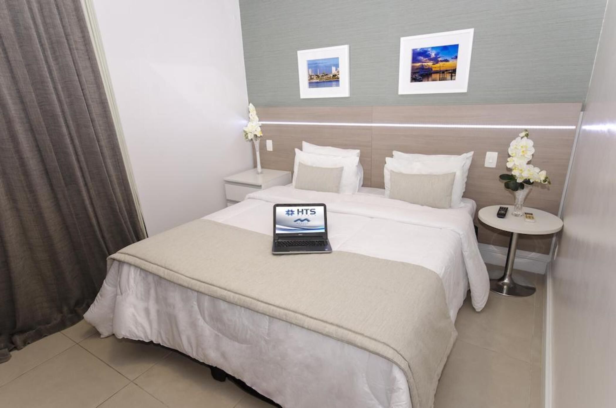A hotel room with a bed, a bedside table with a lamp, a small round table, and two framed pictures on the wall.