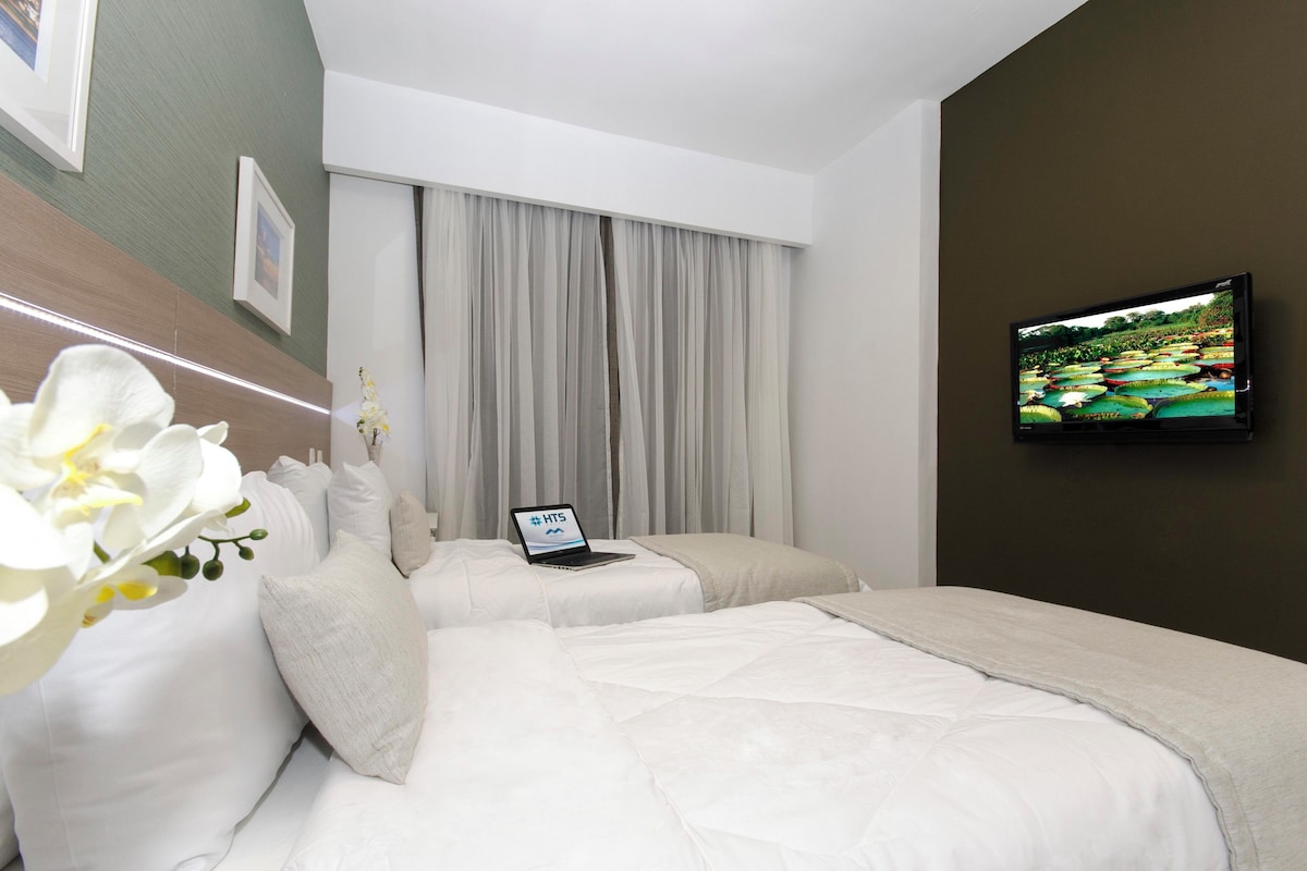 Habitación de hotel con dos camas, una laptop sobre una cama, un televisor en la pared y una ventana con cortinas.
