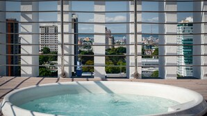 A hot tub on a wooden deck with a cityscape view through a grid-like structure. - Hotel Adrianópolis All Suites (Manaus)