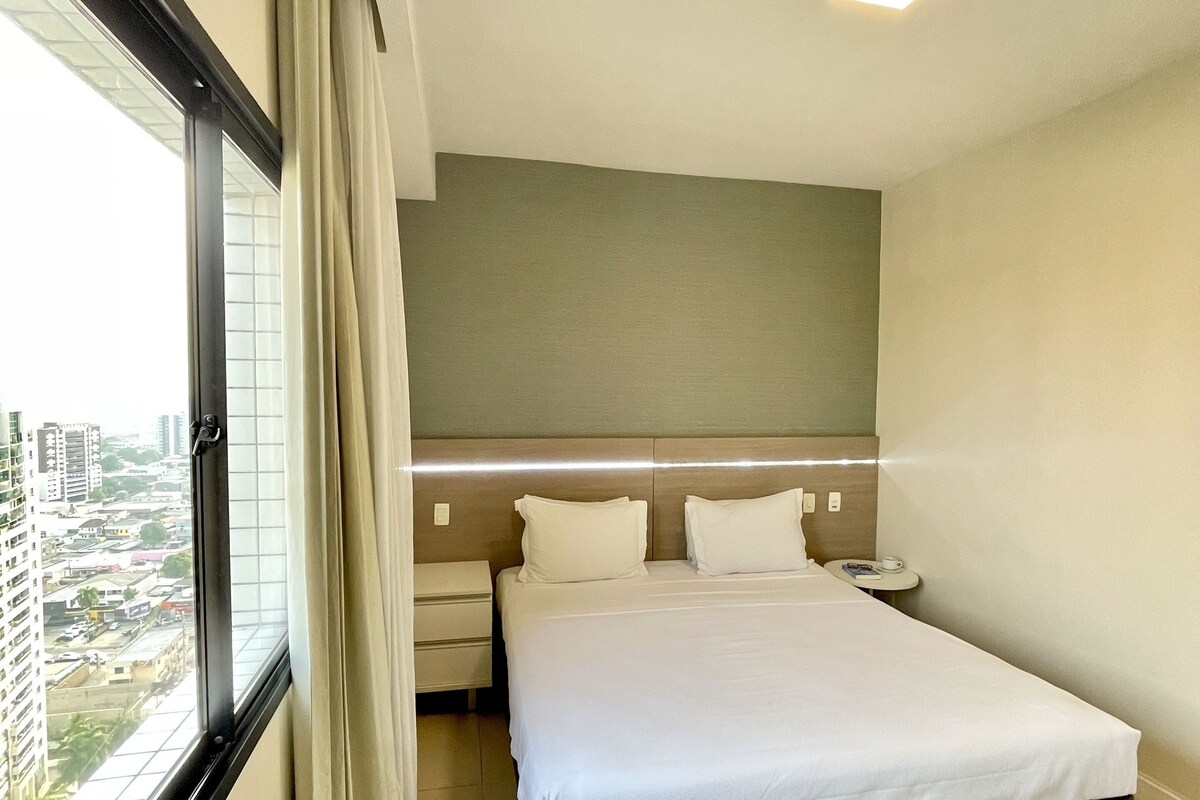 A hotel room with a large bed, a bedside table, a nightstand, a window with curtains, and a city view.
