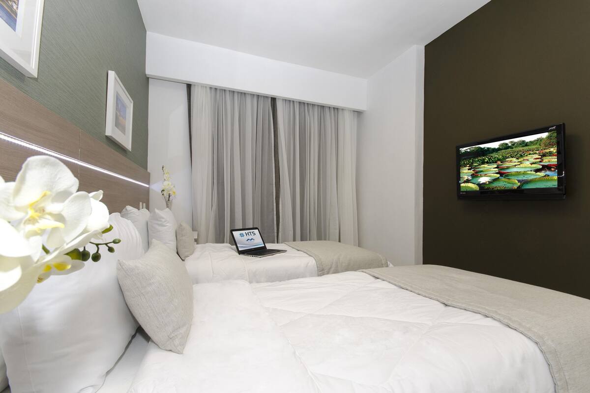 Una camera d'albergo con due letti, un portatile su un letto, una televisione a parete e una finestra con tende.