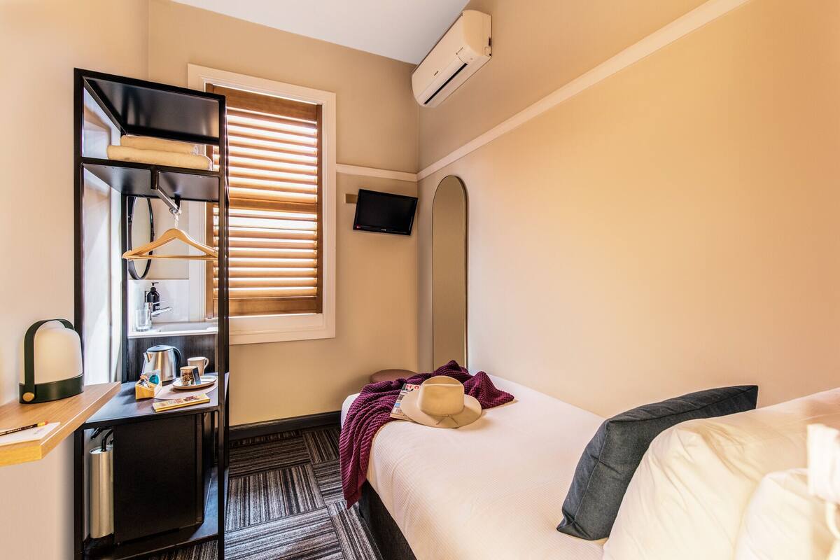 A hotel room with a bed, a desk with a mirror, a hanging light, a window with blinds, and an air conditioning unit.