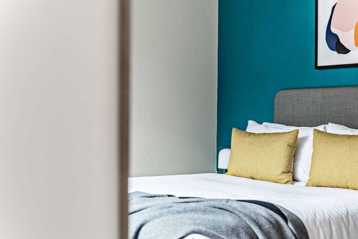 A neatly made bed with white and gray linens, a teal wall, and a framed abstract artwork above it.