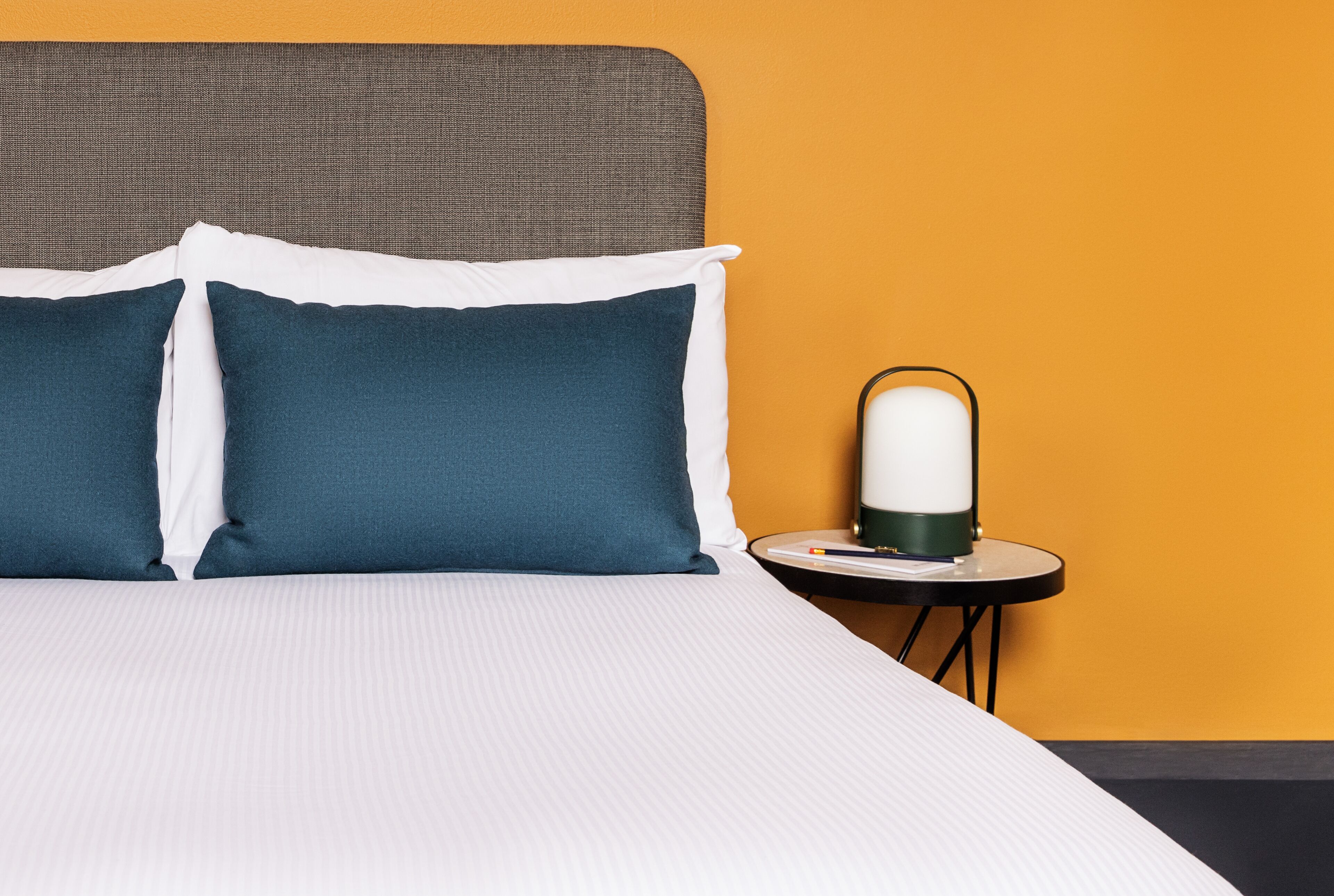 Una cama bien hecha con almohadillas blancas y azul oscuro, un cabecero gris y una mesita de noche con una lámpara.