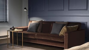 Ein braunes Sofa mit mehreren Kissen, ein kleiner runder Tisch mit einer Lampe und ein Fenster mit Jalousien.