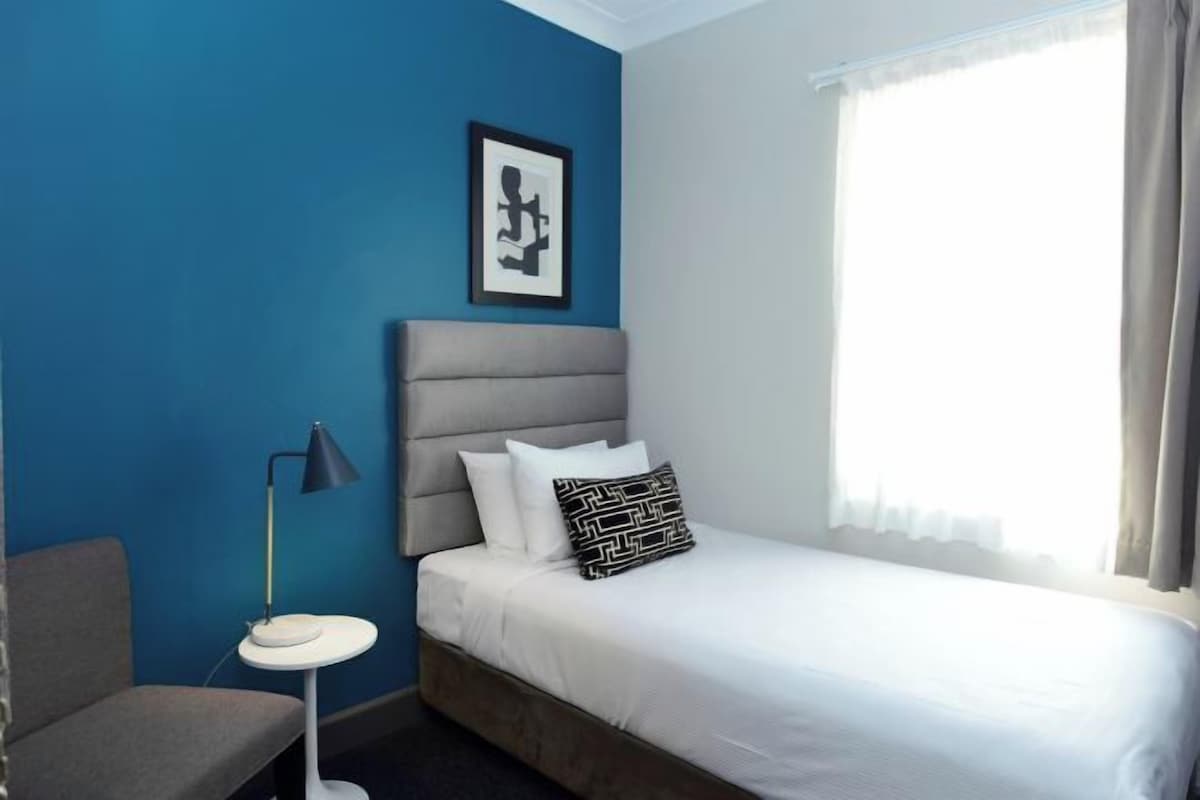 Habitación de hotel con cama, mesita de noche, una silla, lámpara y ventana con cortinas.