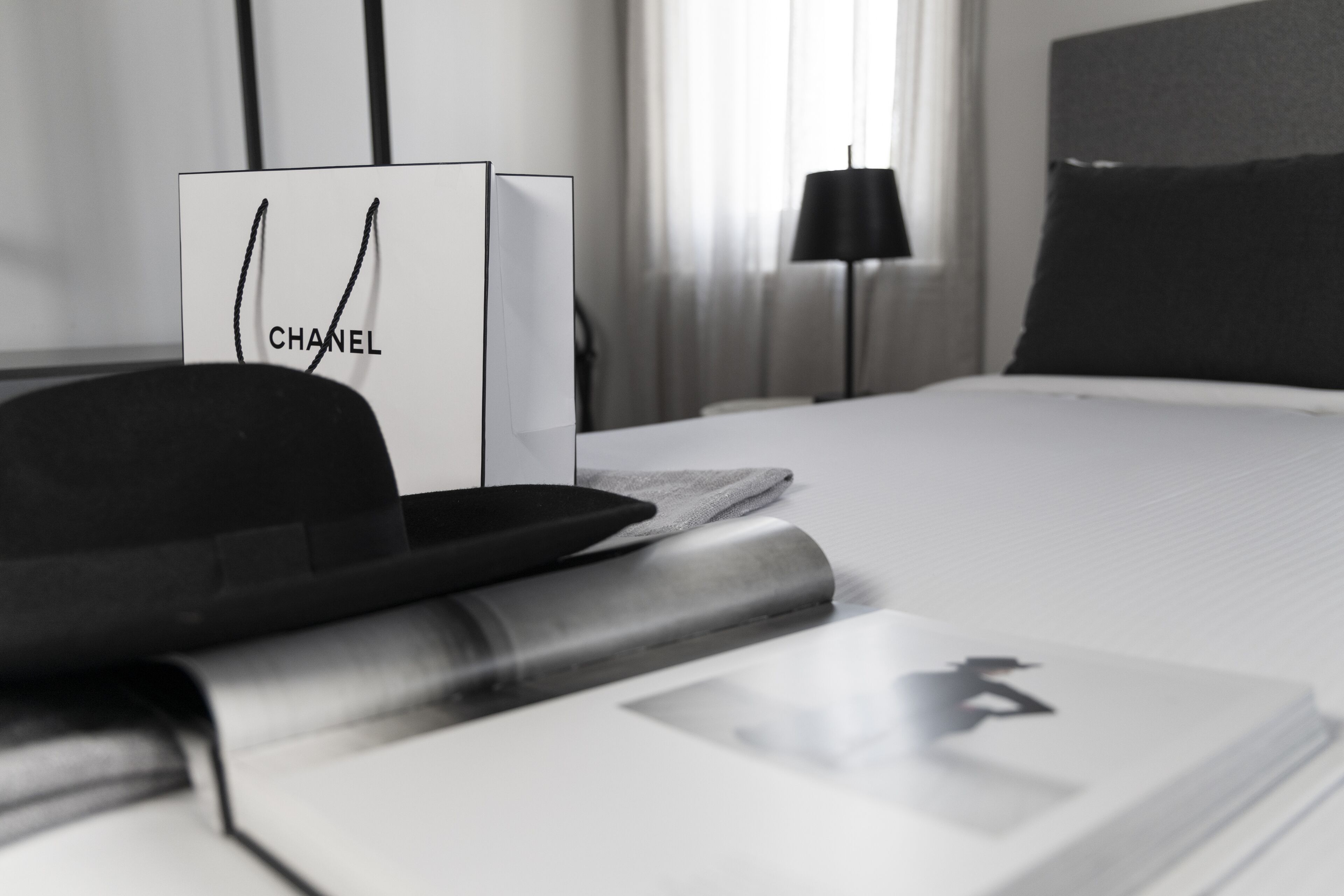 Een net opgemaakt bed met een witte deken en een zwart kussen. Een zwarte hoed en een Chanel boodschappentas liggen op het bed. Een lamp staat op de achtergrond.