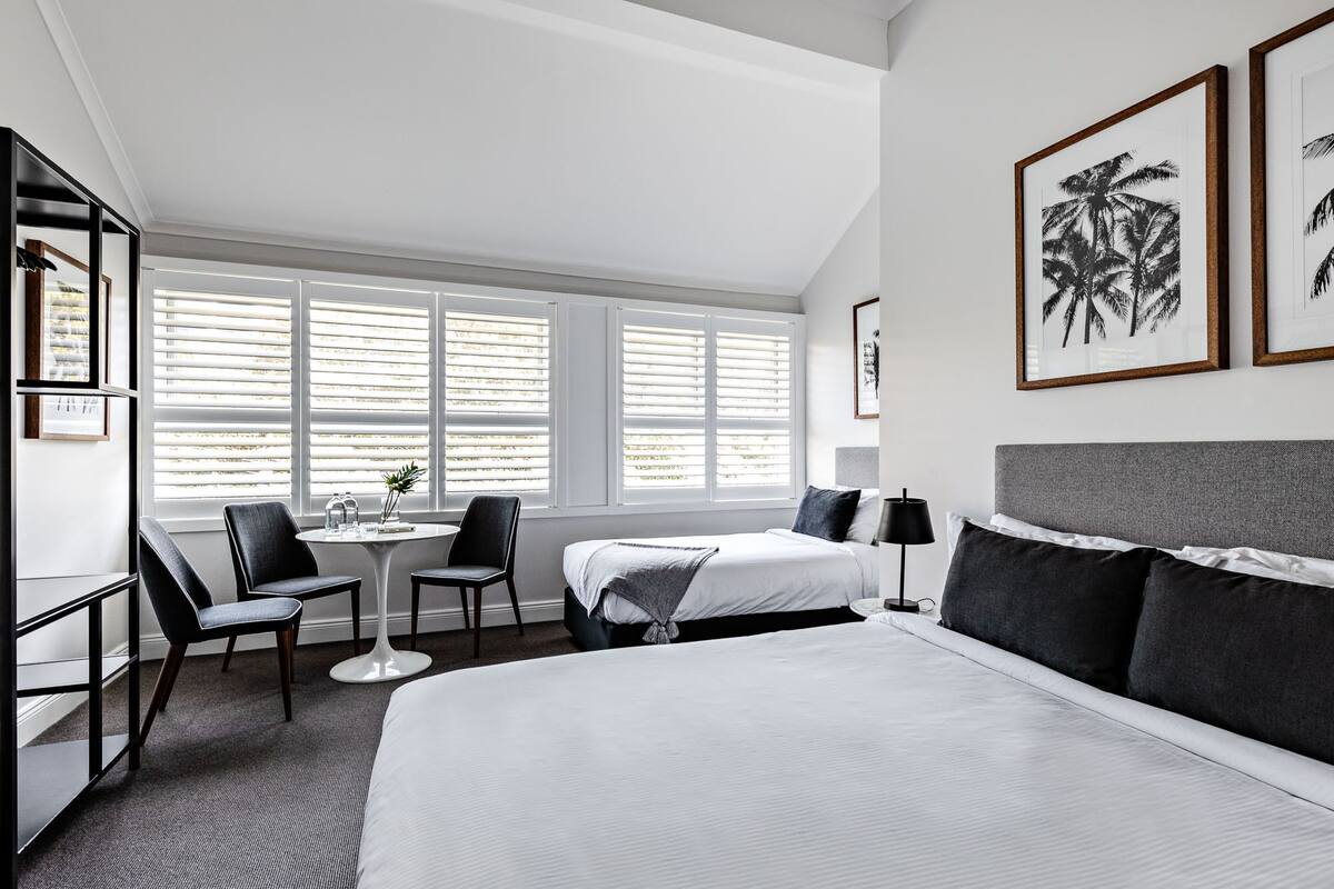 Une chambre d’hôtel avec un grand lit, un bureau, une chaise, une petite table et deux fenêtres équipées de stores.