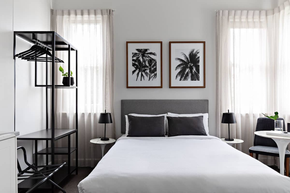 Une chambre d’hôtel moderne avec un grand lit, deux tables de chevet, un bureau avec une chaise, une armoire et deux tableaux encadrés au mur.