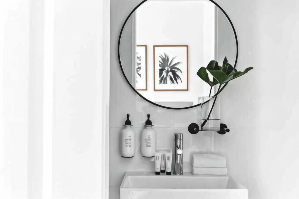 Une salle de bain avec un lavabo blanc, un miroir rond, des œuvres d’art encadrées et une plante en pot.