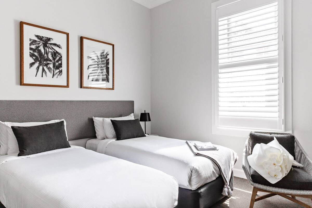 Une chambre d’hôtel avec deux lits, une tête de lit grise, une fenêtre avec des stores et deux tableaux encadrés accrochés au mur.