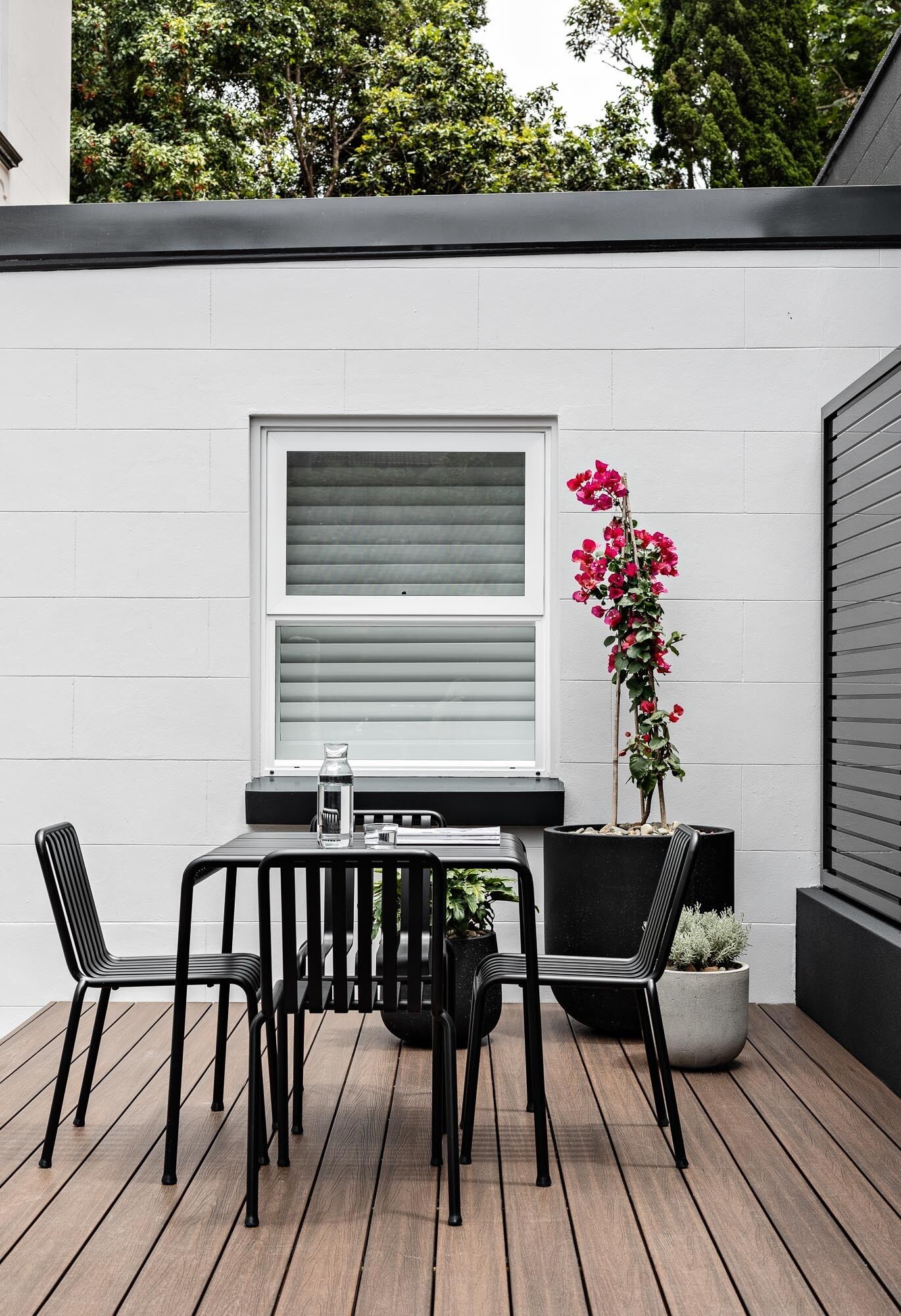 Une terrasse avec une table et des chaises, un pot de fleurs et une fenêtre avec des stores.