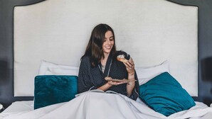 Eine Person sitzt auf einem Bett mit weißem Kopfteil und hält einen Donut.