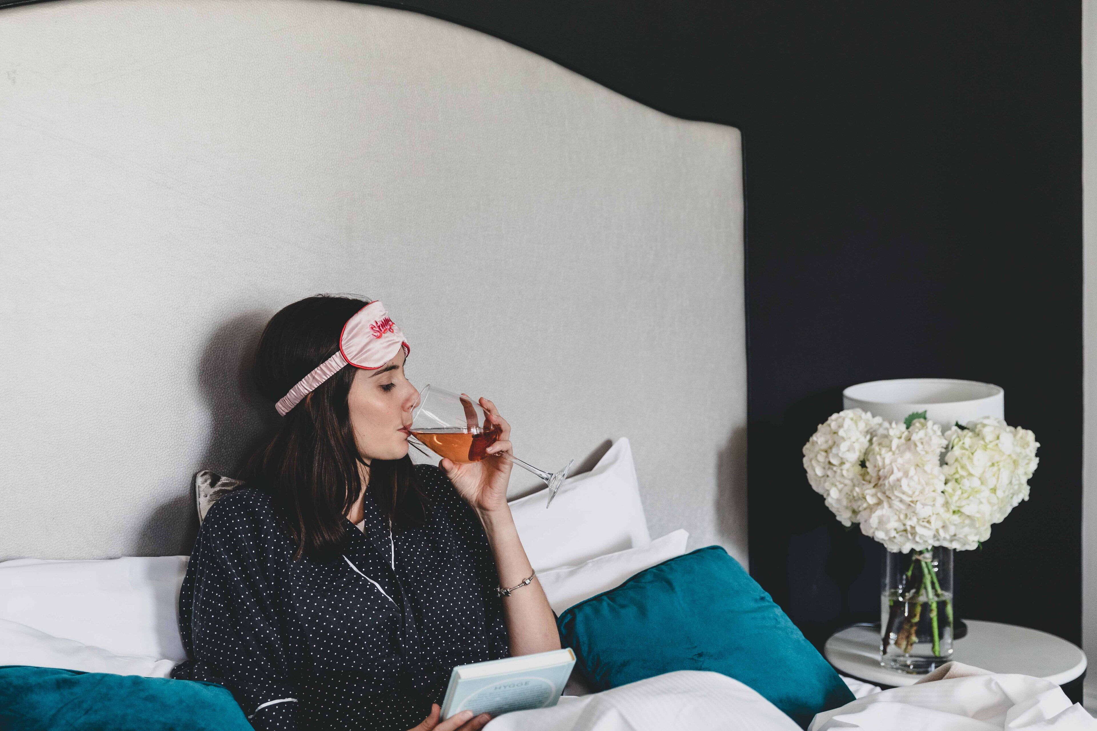 A person sitting on a bed with a book, a glass of drink, and a vase of flowers on a bedside table.