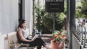 Eine Frau sitzt auf einer Bank und liest ein Buch, im Hintergrund ein Schild mit der Aufschrift 'High Cross'.