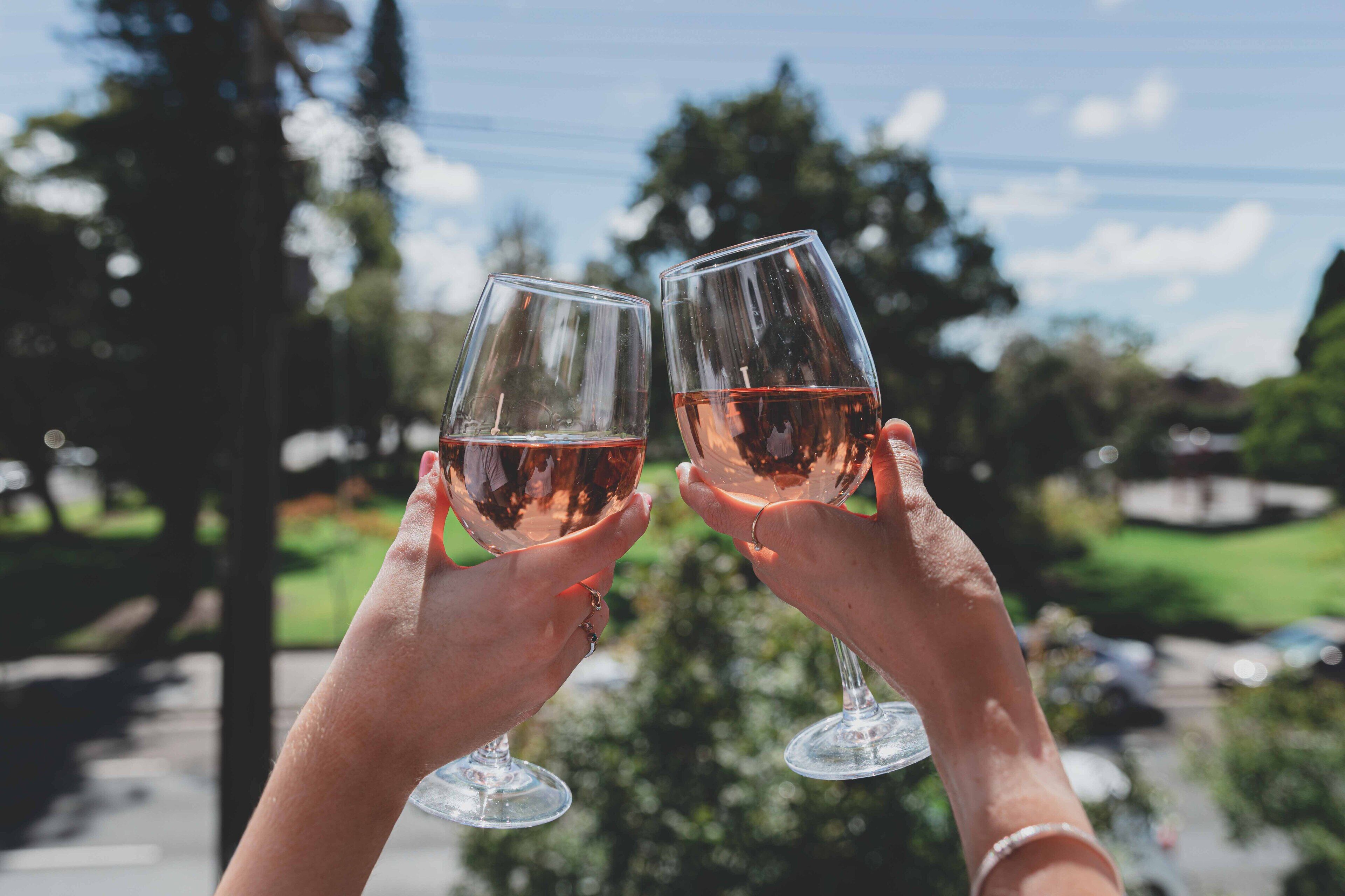 Dos manos sosteniendo copas de vino con un líquido transparente, sobre un fondo de árboles y un cielo despejado.