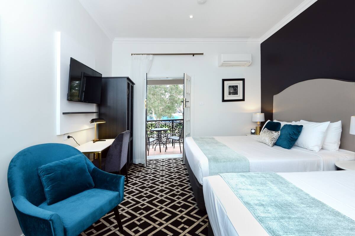 A hotel room with a large bed, a desk, a chair, a TV, and a balcony with outdoor seating.