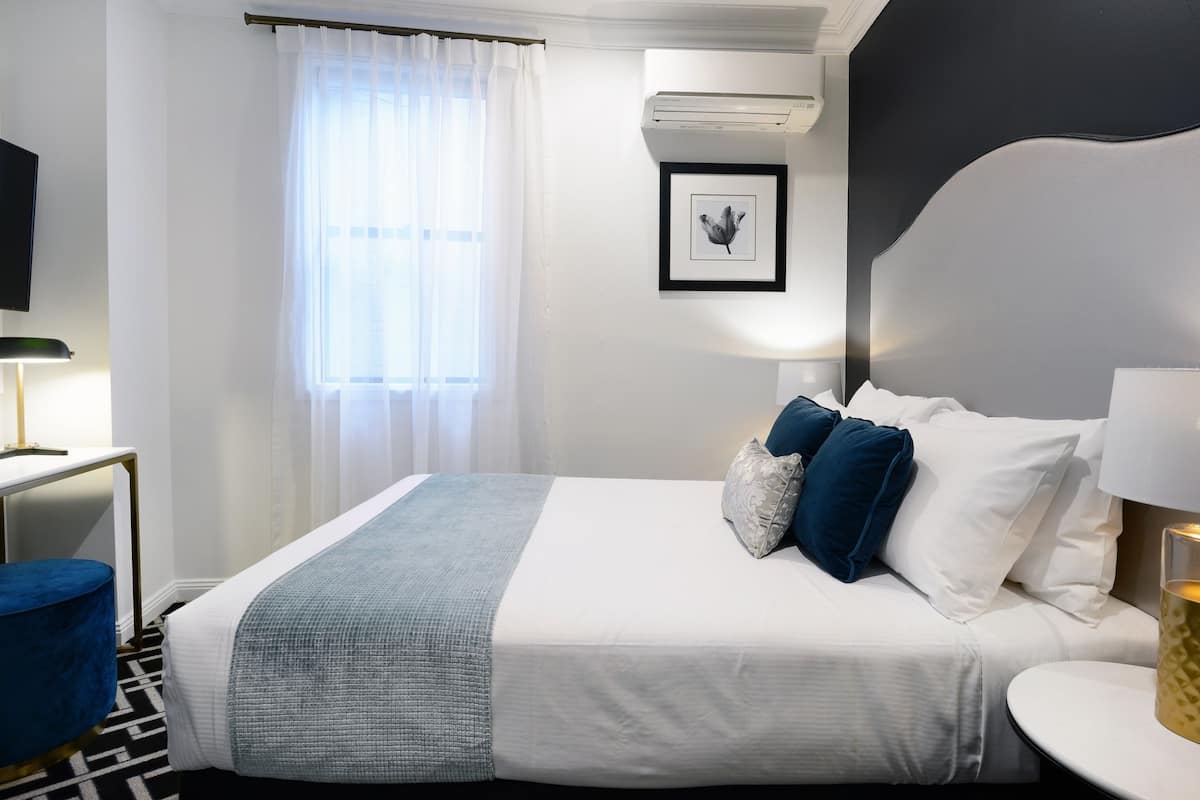 A hotel room with a neatly made bed, a bedside table with a lamp, a blue ottoman, a window with sheer curtains, and a framed picture on the wall.