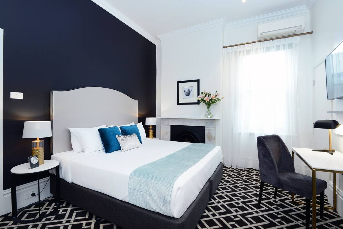 A modern hotel room with a large bed, a small table, a chair, a fireplace, and a TV.