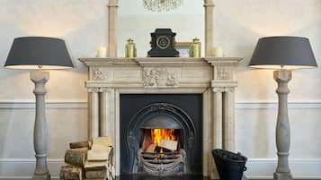 A fireplace with a clock above it, flanked by two lamps, and a stack of books beside it.