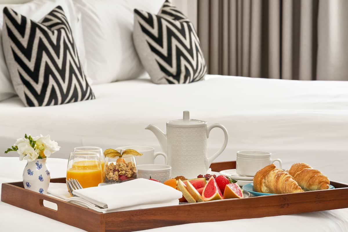 A breakfast tray with croissants, fruit, juice, and coffee on a bed with white bedding and black-and-white patterned pillows.