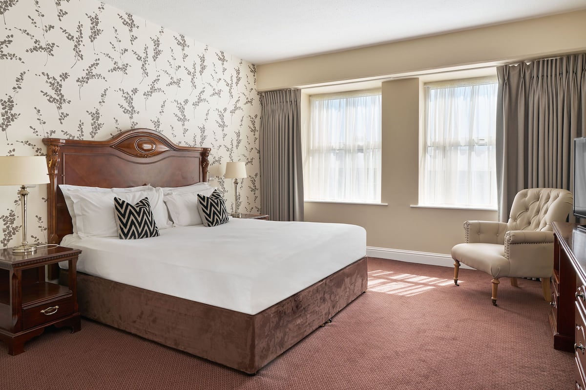 A neatly made bed with a wooden headboard, two bedside lamps, and decorative pillows. A chair is positioned near a window with curtains, and a dresser is visible in the room.