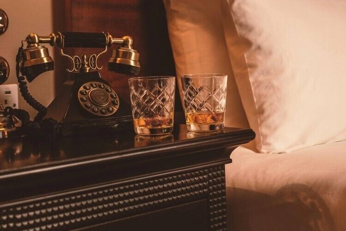 A neatly made bed with white pillows and a wooden headboard. A vintage rotary phone and two crystal glasses are placed on a bedside table.