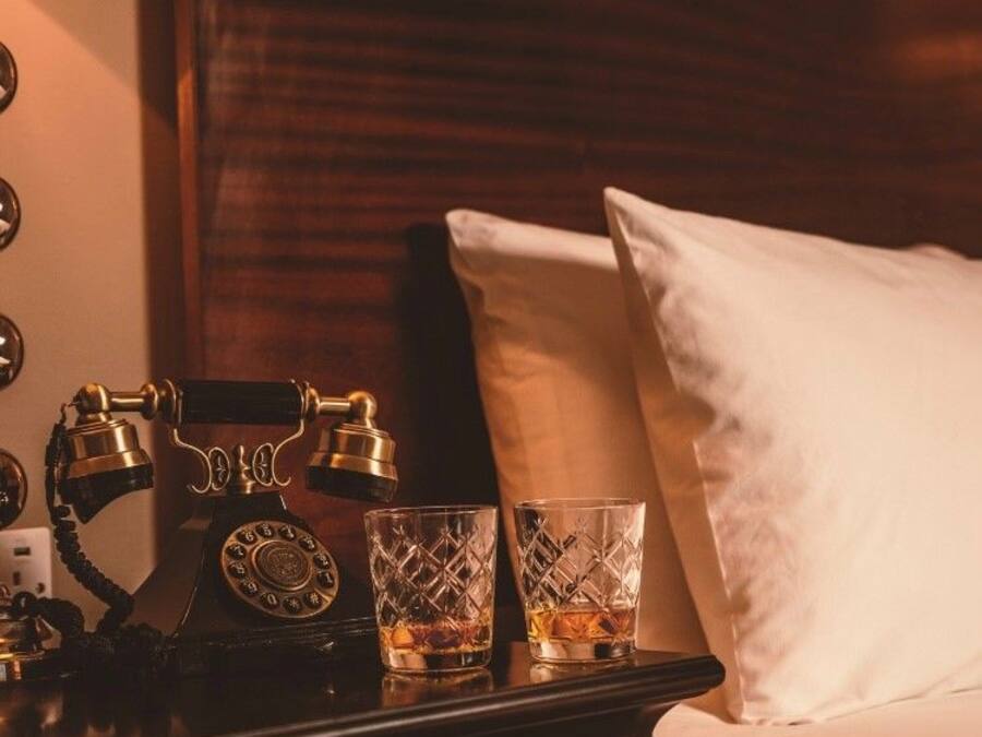 A neatly made bed with white pillows and a wooden headboard. A vintage rotary phone and two crystal glasses are placed on a bedside table.