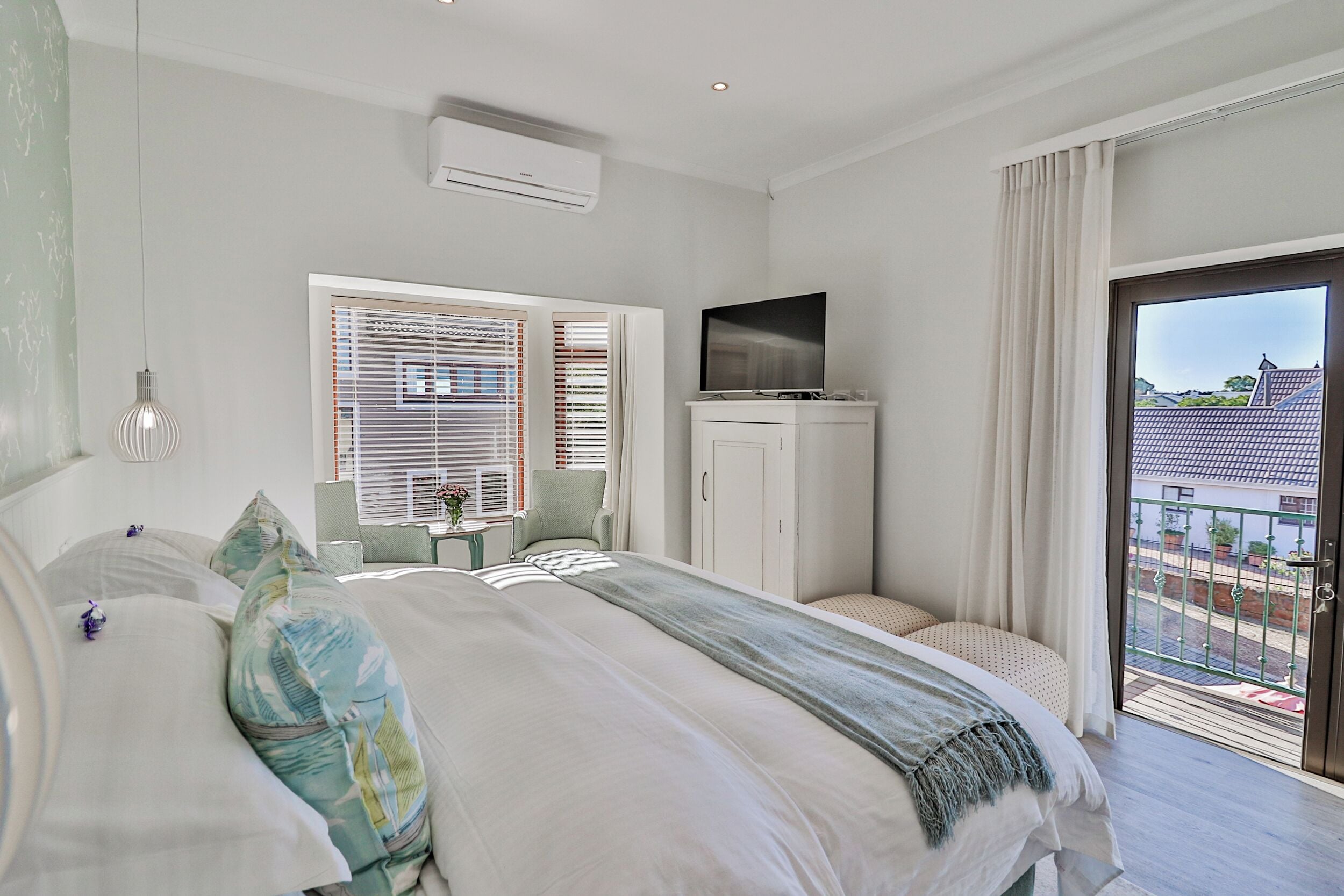 A bedroom with a bed, a TV, a window with blinds, a chair, and a balcony.